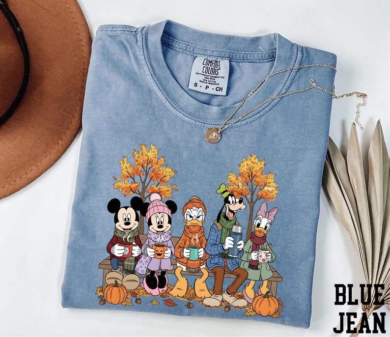 A blue t-shirt with a cartoon image of Disney characters Mickey Mouse, Minnie Mouse, Donald Duck, and Goofy sitting on a bench with autumn leaves and pumpkins.