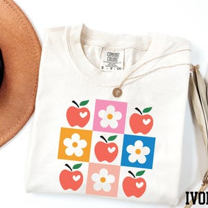 a white t-shirt with a colorful graphic design of apples and flowers, along with a brown hat and a fan.