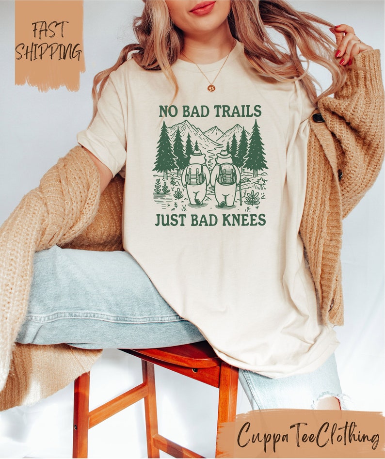 Może przedstawiać: Biały t-shirt z zielonym nadrukiem przedstawiającym dwa niedźwiedzie wędrujące po lesie z tekstem "No Bad Trails Just Bad Knees".