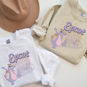May include: Two white sweatshirts with a purple dragon and the text "Epcot" in a retro font. The dragon is standing in front of a drawing of the Epcot Spaceship Earth.