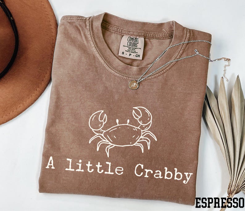 A brown t-shirt with a white crab design and the text "A Little Crabby" printed on it, along with a necklace and a hat.