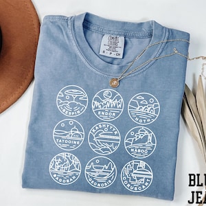 May include: A blue Comfort Colors t-shirt with white graphic circles featuring Star Wars planet designs. The shirt has the words "BLUE JEAN" printed on the bottom right.