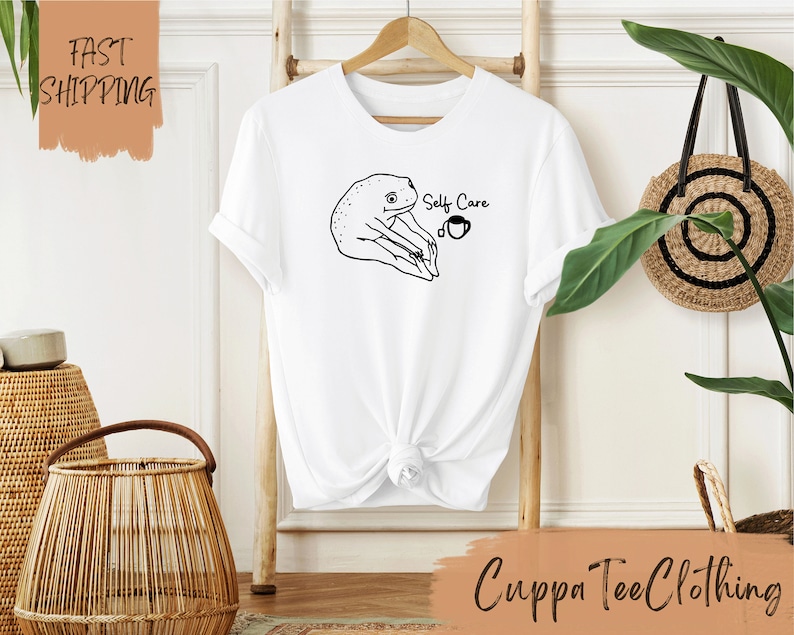 May include: White t-shirt with a black line drawing of a frog sitting with a cup of tea. The text 'Self Care' is printed on the shirt.