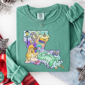 a green long-sleeved shirt with a colorful illustration of Louisiana, a state in the United States, on it. The shirt is laid out on a white surface, along with a pair of jeans and some decorative star-shaped embellishments.