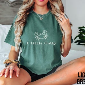 A woman wearing a green t-shirt with a crab design and the text "A little Crabby" is sitting on a bed.