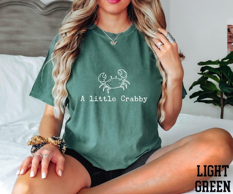 A woman wearing a green t-shirt with a crab design and the text "A little Crabby" is sitting on a bed.