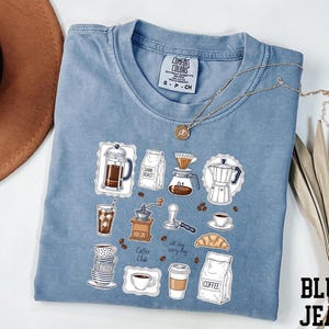 A blue t-shirt with a graphic design of various coffee-related items, such as a coffee maker, coffee cup, and coffee bag, along with the text "Coffee Club" and "All day every day".