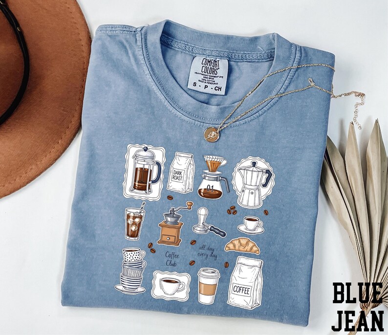 A blue t-shirt with a graphic design of various coffee-related items, such as a coffee maker, coffee cup, and coffee bag, along with the text "Coffee Club" and "All day every day".