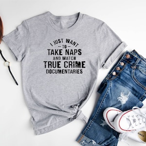 May include: A gray t-shirt with the text "I just want -to- take naps and watch true crime documentaries". The shirt is laid out on a white surface with a pair of jeans, a watch, a camera, and a pair of glasses.