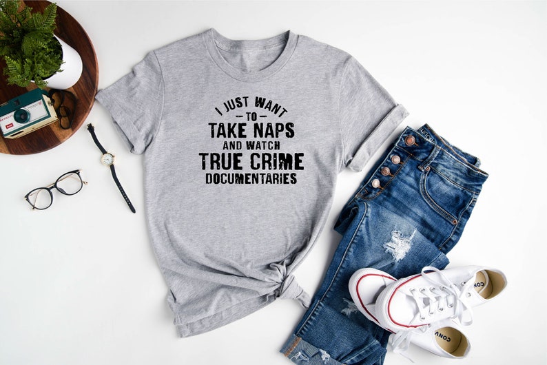 May include: A gray t-shirt with the text "I just want -to- take naps and watch true crime documentaries". The shirt is laid out on a white surface with a pair of jeans, a watch, a camera, and a pair of glasses.