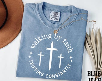 May include: A blue short-sleeved t-shirt featuring a white circular graphic with three crosses and the text "walking by faith TRIPPING CONSTANTLY". The shirt has a crew neckline and a Comfort Colors brand label on the inside collar, size small.