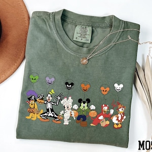 A green t-shirt with a graphic of various Disney characters, including Mickey Mouse, Donald Duck, Goofy, and others, along with balloons. The t-shirt is placed on a white surface, and there is a brown hat and some dried leaves nearby.
