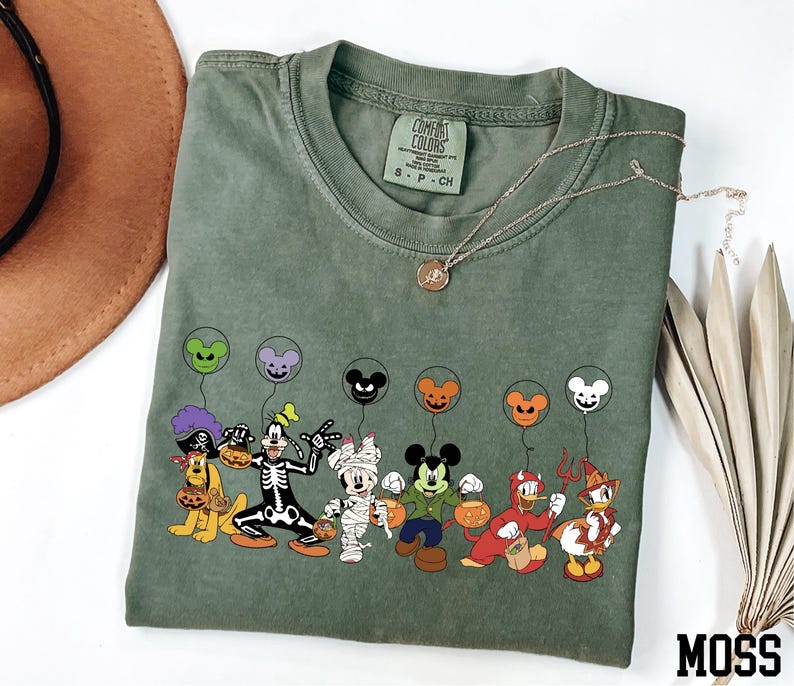 A green t-shirt with a graphic of various Disney characters, including Mickey Mouse, Donald Duck, Goofy, and others, along with balloons. The t-shirt is placed on a white surface, and there is a brown hat and some dried leaves nearby.