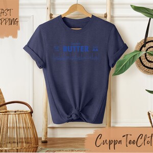 Puede incluir: Una camiseta azul marino con un gr&aacute;fico azul que dice "Salted Butter" con un gr&aacute;fico de escala debajo. La camiseta est&aacute; en una percha de madera frente a una pared blanca.