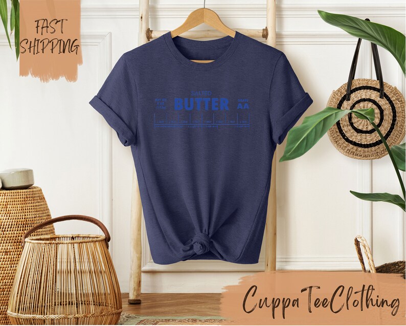 Puede incluir: Una camiseta azul marino con un gr&aacute;fico azul que dice "Salted Butter" con un gr&aacute;fico de escala debajo. La camiseta est&aacute; en una percha de madera frente a una pared blanca.