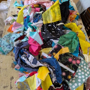 May include: A collection of colourful fabric scraps in various sizes and patterns, including floral, polka dot, and abstract designs. The fabrics are in shades of yellow, pink, blue, and black, suggesting a quilting or sewing project.