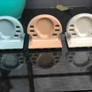 May include: Three ceramic toothbrush holders in pastel shades: light blue, beige, and speckled white. Each holder features a rounded top with holes for toothbrushes and a square base. The holders are displayed on a reflective surface.