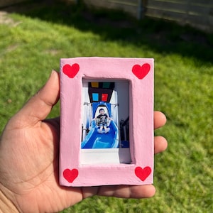 May include: Pink wooden picture frame with red hearts around the edges. The frame holds a photo of a child on a blue slide.