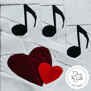 May include: A white fabric quilt block with three black musical notes and two red hearts. The quilt block has a white background with a pattern of white fabric squares sewn together. The musical notes are in the upper part of the block and the hearts are in the lower part of the block. The quilt block has a white circle with the text "Chris & Dell PATTERNS" in black.