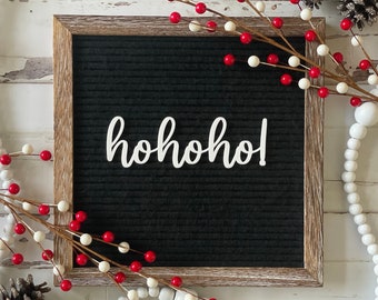 Può includere: Una lavagna nera con una cornice in legno bianco e il testo "ho ho ho!" scritto su di essa. La lavagna è circondata da rametti di bacche bianche e rosse e perline bianche.