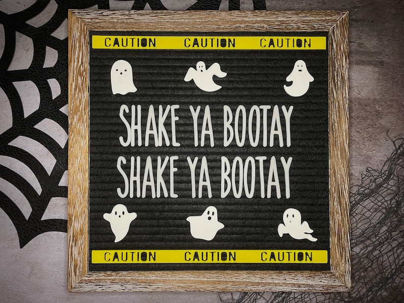 May include: A black letter board with the text "Shake Ya Bootay" written in white letters. The board is surrounded by a wooden frame and has yellow caution tape at the top and bottom. There are six white ghost silhouettes on the board.