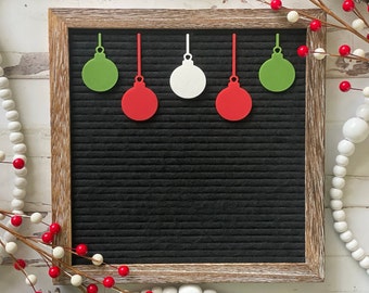 Può includere: Una lavagna nera con cornice in legno, decorata con cinque ornamenti di carta. Gli ornamenti sono rossi, verdi e bianchi e hanno la forma di addobbi per l'albero di Natale.