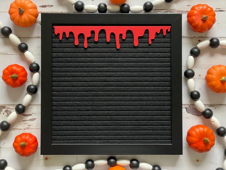 K&ouml;nnte beinhalten: Ein schwarzes Buchstabenbrett mit einem roten, tropfenden Blutsaum. Das Brett ist von wei&szlig;en und schwarzen Perlen und orangefarbenen K&uuml;rbissen umgeben.