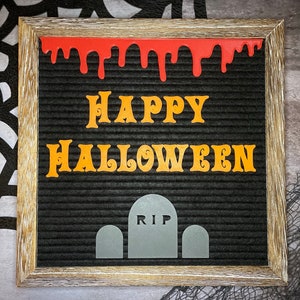 K&ouml;nnte beinhalten: Ein schwarzes Buchstabenbrett mit einem Holzrahmen. Das Brett zeigt "Happy Halloween" in orangefarbenen Buchstaben mit roter, tropfender Farbe &uuml;ber den Buchstaben. Am unteren Rand des Bretts befinden sich drei graue Grabsteine, wobei der mittlere Grabstein "RIP" sagt.
