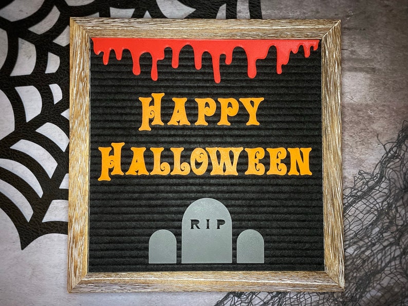 K&ouml;nnte beinhalten: Ein schwarzes Buchstabenbrett mit einem Holzrahmen. Das Brett zeigt "Happy Halloween" in orangefarbenen Buchstaben mit roter, tropfender Farbe &uuml;ber den Buchstaben. Am unteren Rand des Bretts befinden sich drei graue Grabsteine, wobei der mittlere Grabstein "RIP" sagt.