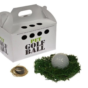 May include: A white cardboard box with a handle and the words "Pet Golf Ball" printed on it. The box contains a white golf ball and a bed of green grass.