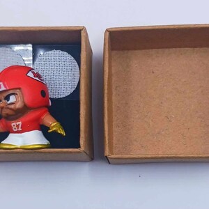 May include: A small, red and white football player figurine in a brown cardboard box. The figurine is wearing a helmet with the word "CHIEFS" on it and the number "87" on its jersey.