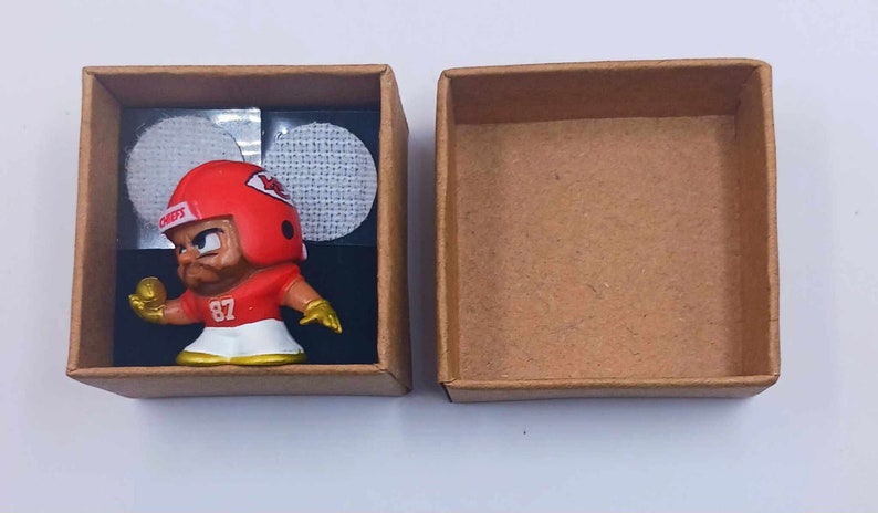 May include: A small, red and white football player figurine in a brown cardboard box. The figurine is wearing a helmet with the word "CHIEFS" on it and the number "87" on its jersey.