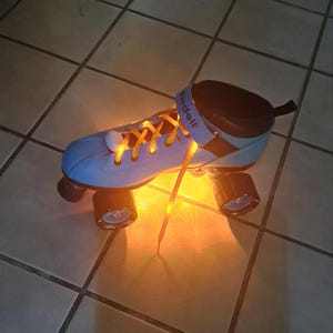 Puede incluir: Un patín de ruedas azul claro con cordones naranjas y luces naranjas brillantes. El patín tiene ruedas negras y un cuello acolchado negro y marrón. La marca "Riedell" es visible en la correa.