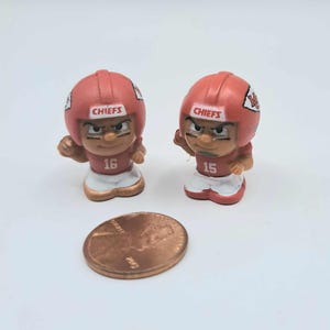 May include: Two small plastic figurines of football players wearing red and white uniforms with the word "CHIEFS" on their helmets. The players are wearing the numbers 16 and 15 on their jerseys. A penny is included for scale.