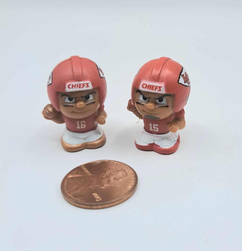 May include: Two small plastic figurines of football players wearing red and white uniforms with the word "CHIEFS" on their helmets. The players are wearing the numbers 16 and 15 on their jerseys. A penny is included for scale.