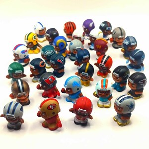 May include: A collection of miniature football players wearing different team uniforms. The players are made of plastic and have detailed features, including helmets, jerseys, and pants. The players are arranged in a circle on a white background.