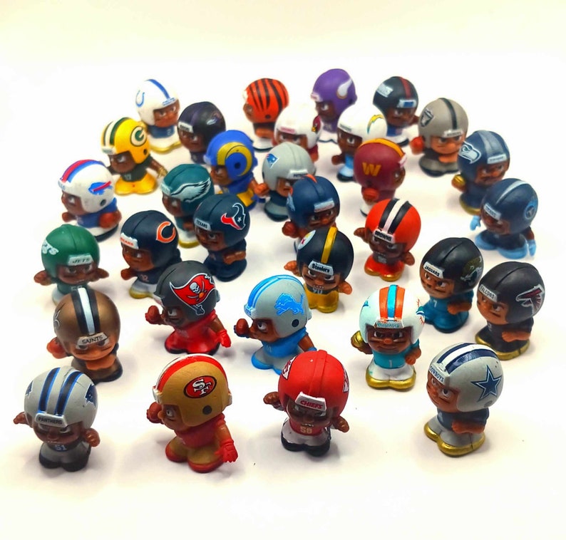 May include: A collection of miniature football players wearing different team uniforms. The players are made of plastic and have detailed features, including helmets, jerseys, and pants. The players are arranged in a circle on a white background.