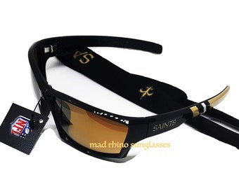 May include: Black sunglasses with brown lenses and a black strap with gold lettering that says "Saints". The strap has a gold fleur-de-lis logo. The sunglasses are labeled "mad rhino sunglasses".