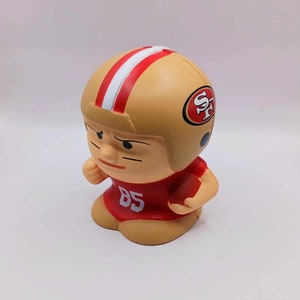 Puede incluir: Figura de un jugador de fútbol americano con un casco dorado con rayas rojas y blancas y el logo de los 49ers de San Francisco. El jugador lleva una camiseta roja con el número 85.