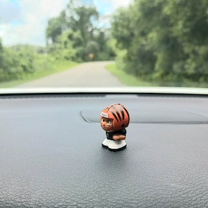 May include: A small plastic figurine of a football player wearing a black and orange uniform with a helmet that says "Bengals". The figurine is sitting on a black surface.