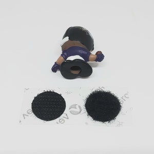 May include: A small plastic figure of a football player wearing a purple and black uniform. The figure is holding a football and has a black helmet with a white logo. The figure is attached to a piece of black hook and loop fastener.