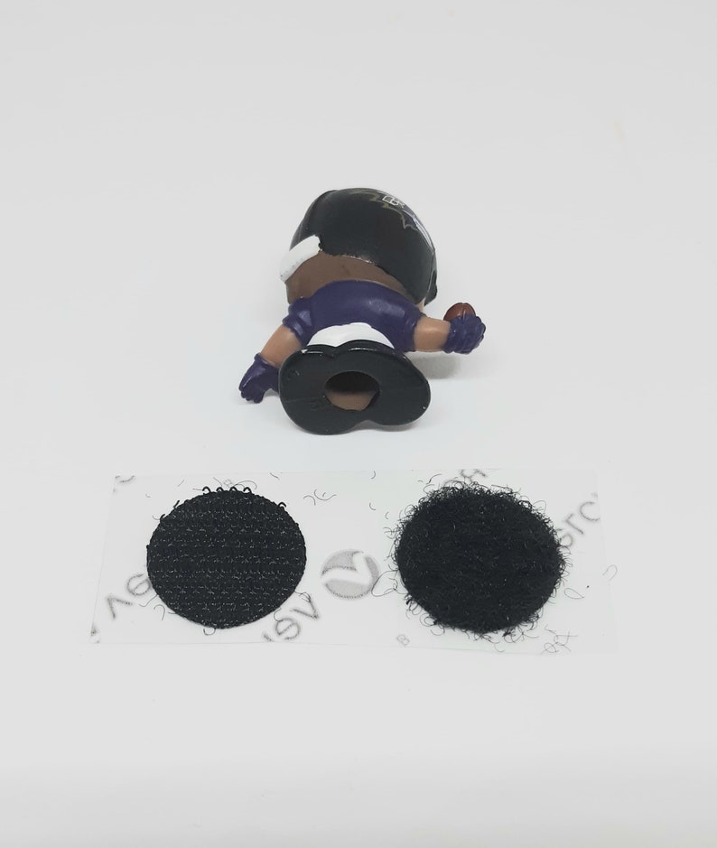 May include: A small plastic figure of a football player wearing a purple and black uniform. The figure is holding a football and has a black helmet with a white logo. The figure is attached to a piece of black hook and loop fastener.