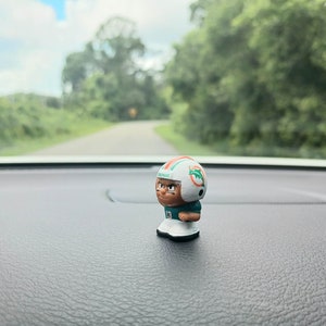 Peut inclure: Une petite figurine en plastique d'un joueur de football américain portant un casque et un maillot vert et blanc avec le mot "Dolphins" dessus. La figurine est debout sur une surface noire.