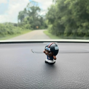May include: A small plastic figurine of a football player wearing a blue and white uniform with a helmet that says "Giants" on it. The figurine is standing on a black surface.