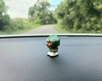 May include: A green and black figurine of a football player wearing a helmet with a white wing logo. The figurine is standing on a black surface.