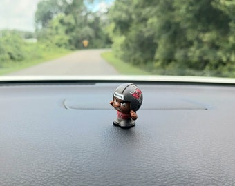 May include: A small black and red football player figurine with a helmet and a determined expression. The figurine is sitting on a black dashboard.