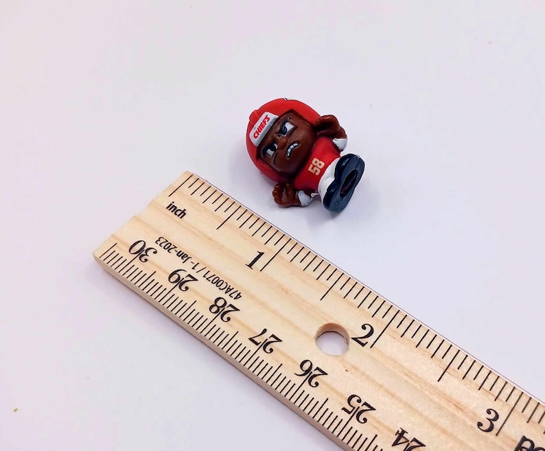 May include: A small red and black plastic figurine of a football player wearing a helmet with the word "CHIEFS" on it. The player is wearing a jersey with the number "58" on it. The figurine is about 1 inch tall.