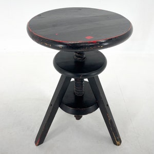 May include: A black wooden adjustable stool with three legs and a round seat. The stool has a red painted edge and a worn finish.