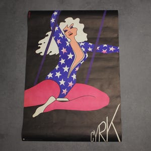 May include: A vintage circus poster featuring a woman in a blue and white star-patterned leotard and pink tights, swinging on a trapeze. The poster has a black background and the word "CYRK" is written in white at the bottom.
