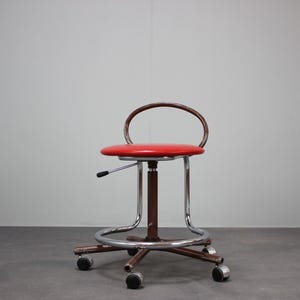 May include: A vintage red and chrome adjustable stool with a circular seat and a curved backrest. The stool has a brown central column, a chrome base, and four rolling wheels. The height adjustment lever is visible.